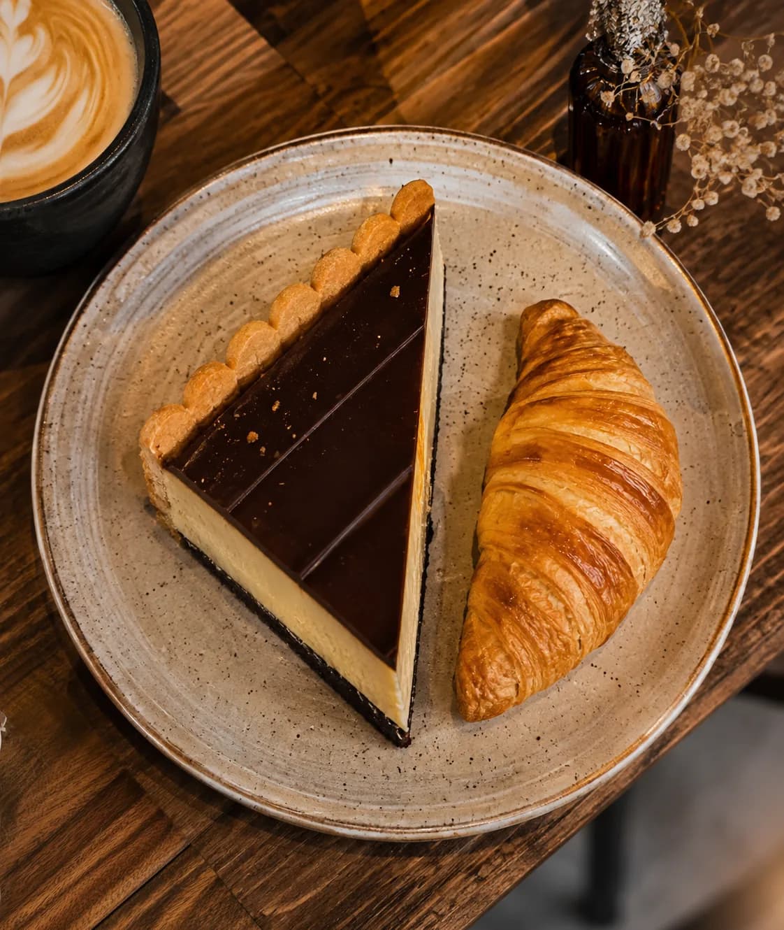 Fatia de torta de chocolate e croissant servidos em prato de cerâmica sobre mesa de madeira.